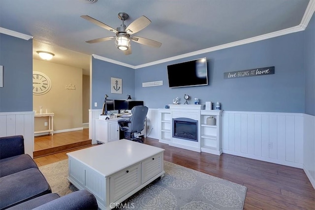 living room featuring a wainscoted wall, ornamental molding, a glass covered fireplace, ceiling fan, and wood finished floors