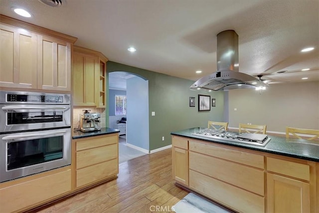 kitchen featuring arched walkways, light brown cabinets, island range hood, stainless steel appliances, and dark countertops