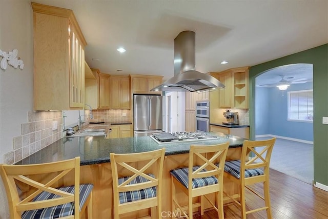 kitchen with arched walkways, appliances with stainless steel finishes, island exhaust hood, light brown cabinetry, and a sink