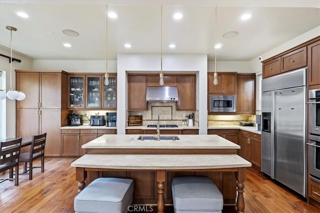 kitchen with a kitchen bar, wood-type flooring, built in appliances, hanging light fixtures, and range hood