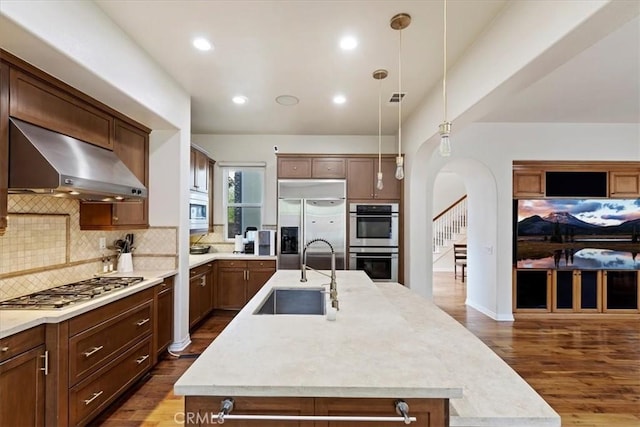 kitchen featuring sink, hanging light fixtures, built in appliances, a kitchen island with sink, and exhaust hood