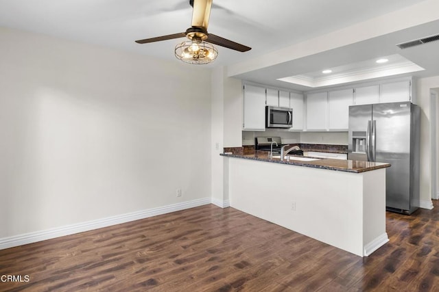 kitchen with kitchen peninsula, appliances with stainless steel finishes, dark hardwood / wood-style flooring, dark stone counters, and a tray ceiling