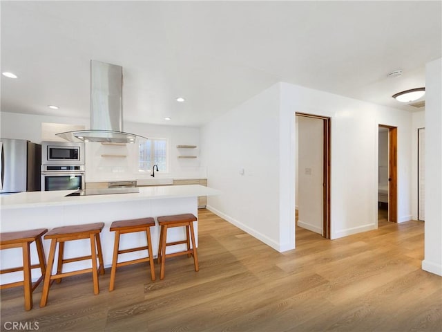 kitchen with a breakfast bar area, appliances with stainless steel finishes, island range hood, kitchen peninsula, and light wood-type flooring