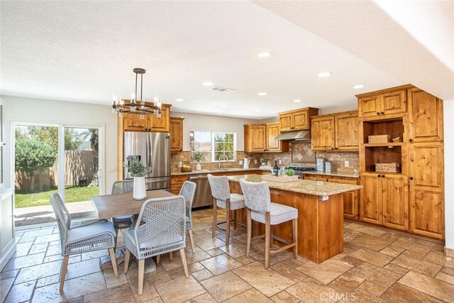 kitchen featuring light stone countertops, backsplash, stainless steel appliances, pendant lighting, and an island with sink