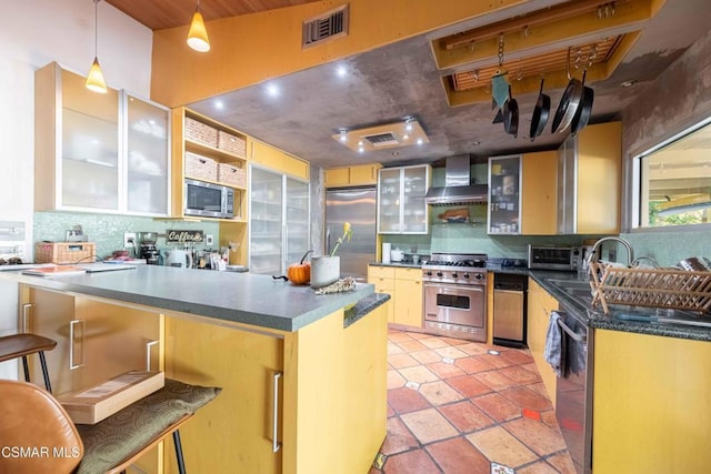 kitchen with kitchen peninsula, hanging light fixtures, built in appliances, wall chimney exhaust hood, and a breakfast bar