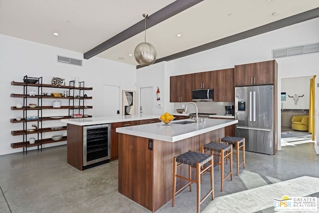 kitchen featuring stainless steel appliances, beverage cooler, decorative light fixtures, a center island with sink, and concrete floors