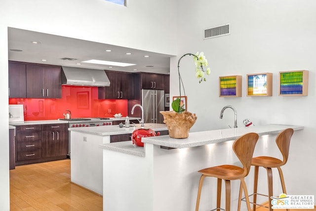 kitchen featuring wall chimney exhaust hood, light hardwood / wood-style flooring, kitchen peninsula, a breakfast bar area, and appliances with stainless steel finishes