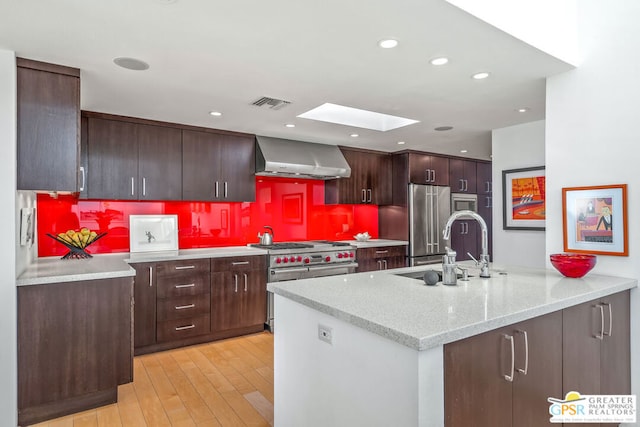 kitchen with a skylight, high end appliances, sink, wall chimney range hood, and light hardwood / wood-style flooring