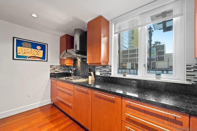 kitchen featuring wall chimney range hood, dark stone countertops, wood-type flooring, stainless steel gas stovetop, and decorative backsplash