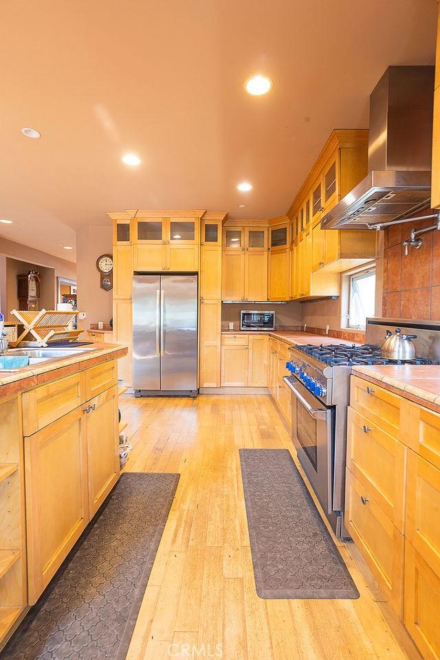 kitchen featuring wall chimney range hood, light countertops, stainless steel appliances, light wood-style floors, and glass insert cabinets