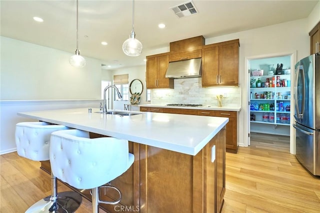 kitchen with an island with sink, pendant lighting, exhaust hood, and stainless steel refrigerator