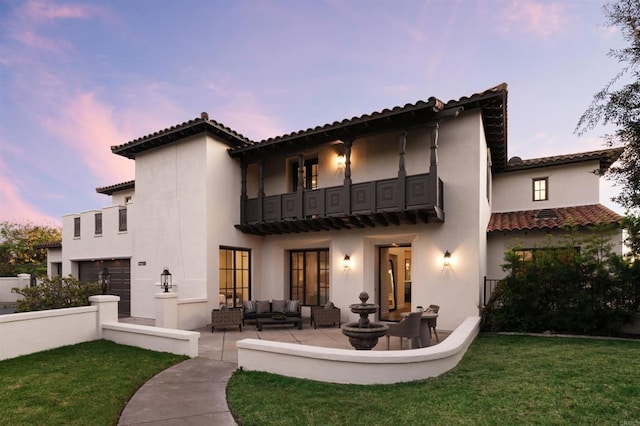 back house at dusk featuring a patio, a balcony, an outdoor hangout area, and a lawn