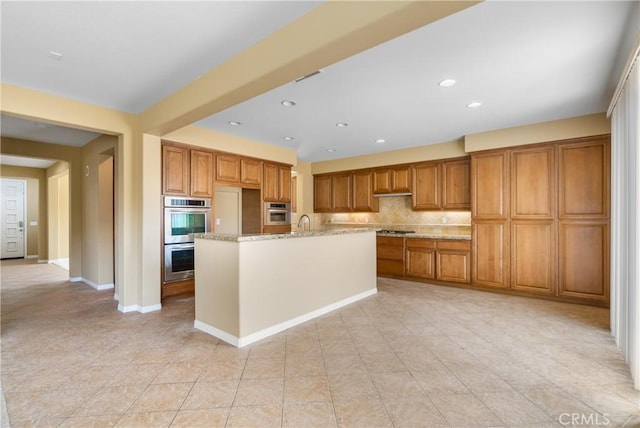 kitchen featuring stainless steel double oven, brown cabinets, light stone countertops, and a kitchen island with sink