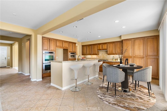 kitchen featuring brown cabinetry, an island with sink, a kitchen breakfast bar, light stone countertops, and double oven