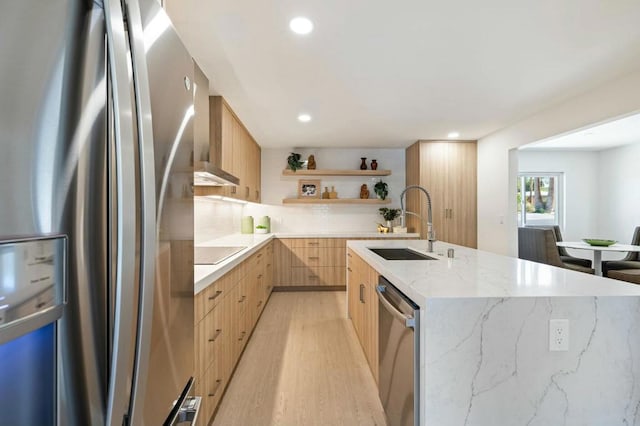 kitchen featuring wall chimney range hood, sink, an island with sink, appliances with stainless steel finishes, and light hardwood / wood-style floors