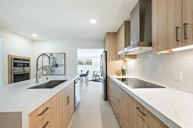 kitchen featuring light stone countertops, appliances with stainless steel finishes, light wood-type flooring, wall chimney exhaust hood, and sink