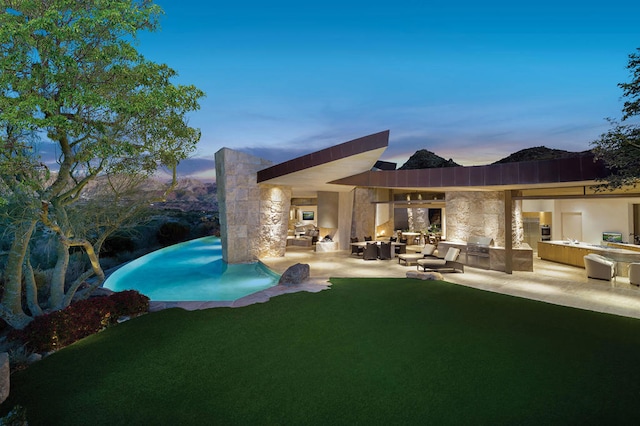 back house at dusk with a lawn, a patio area, an outdoor living space with a fireplace, and area for grilling