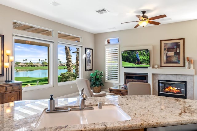 kitchen featuring a water view, sink, ceiling fan, light stone counters, and a tiled fireplace