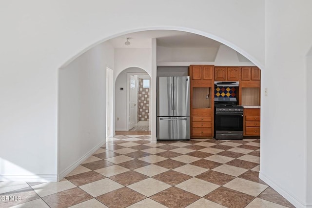 kitchen featuring exhaust hood, range with gas stovetop, freestanding refrigerator, decorative backsplash, and brown cabinetry