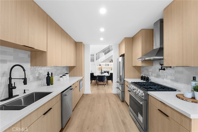 kitchen featuring high end appliances, wall chimney range hood, sink, light hardwood / wood-style flooring, and tasteful backsplash