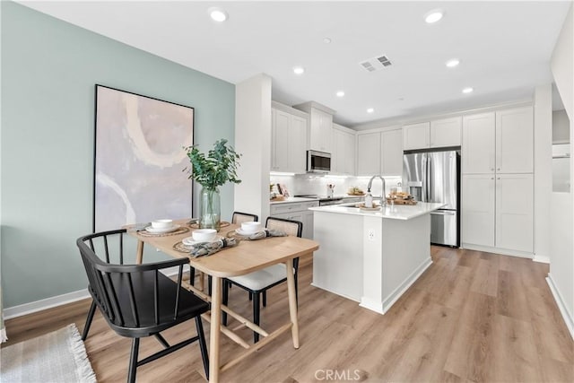 kitchen with appliances with stainless steel finishes, a kitchen island with sink, sink, white cabinets, and light hardwood / wood-style floors