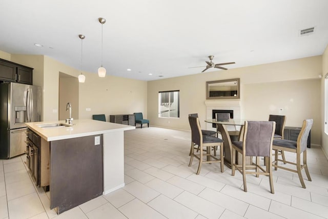 kitchen featuring stainless steel fridge, ceiling fan, a kitchen island with sink, sink, and pendant lighting