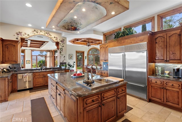 kitchen with sink, stainless steel appliances, backsplash, dark stone counters, and a kitchen island with sink