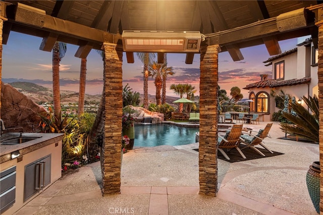 pool at dusk with a gazebo, a patio, exterior kitchen, and sink