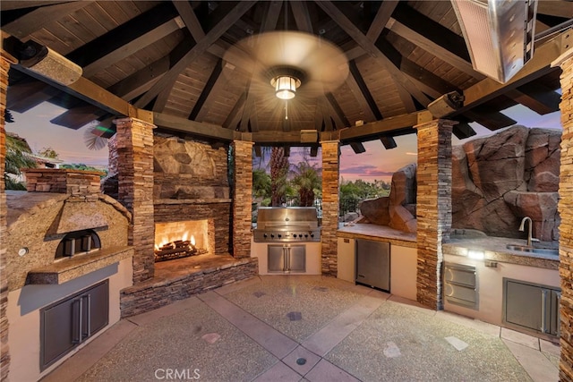 patio terrace at dusk with a gazebo, an outdoor stone fireplace, grilling area, and sink