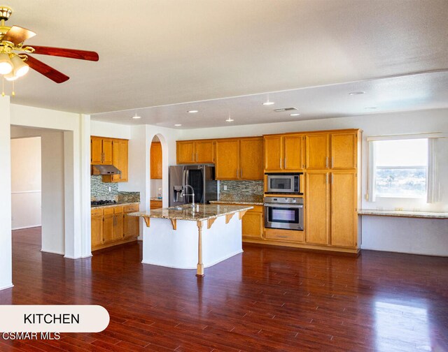 kitchen featuring light stone counters, stainless steel appliances, dark hardwood / wood-style floors, a breakfast bar area, and an island with sink