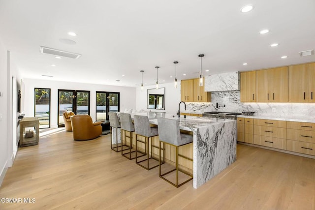 kitchen with light stone countertops, light brown cabinets, wall chimney range hood, a large island with sink, and light wood-type flooring