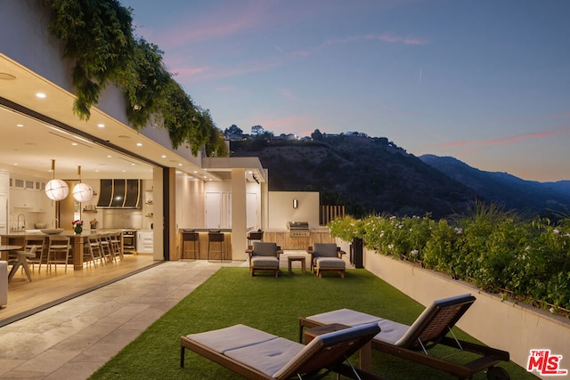 patio terrace at dusk with a lawn, sink, exterior kitchen, a mountain view, and a grill