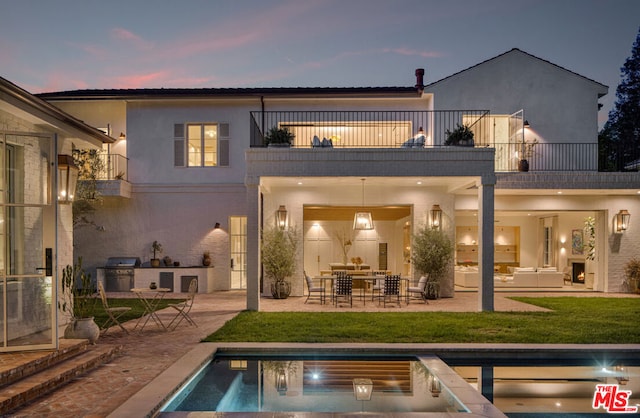 back house at dusk featuring an outdoor kitchen, a balcony, a patio area, and a lawn