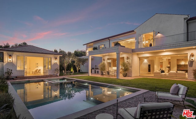 back house at dusk featuring outdoor lounge area, a yard, a pool with hot tub, a balcony, and a patio