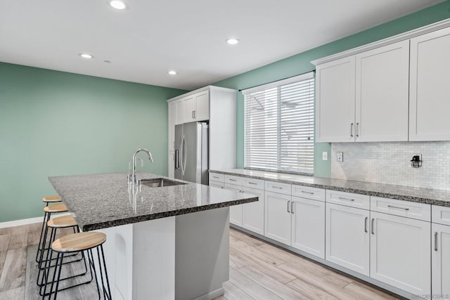 kitchen featuring sink, white cabinetry, a kitchen island with sink, stainless steel refrigerator with ice dispenser, and dark stone counters