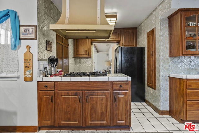 kitchen featuring tile countertops, stainless steel gas stovetop, island range hood, black refrigerator, and light tile patterned flooring