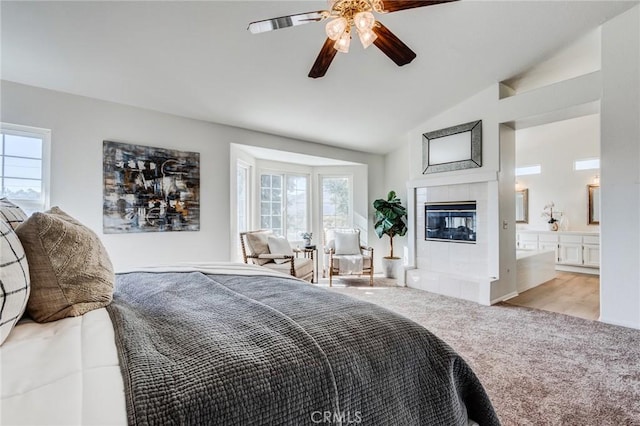 bedroom with ceiling fan, light colored carpet, vaulted ceiling, a tiled fireplace, and ensuite bath