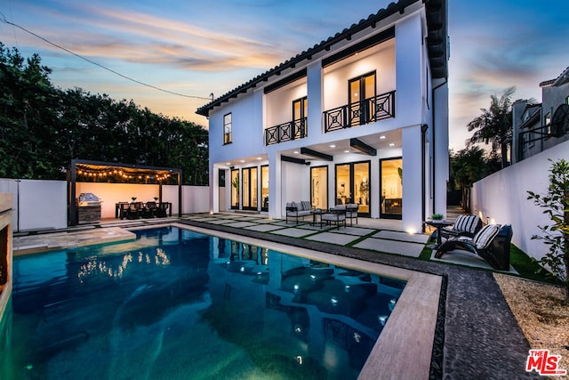 back house at dusk featuring outdoor lounge area, a balcony, a patio area, and a fenced in pool