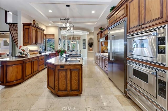 kitchen with a tray ceiling, sink, built in appliances, hanging light fixtures, and an island with sink