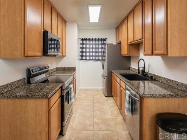 kitchen featuring a sink, dark stone countertops, light tile patterned flooring, brown cabinetry, and appliances with stainless steel finishes