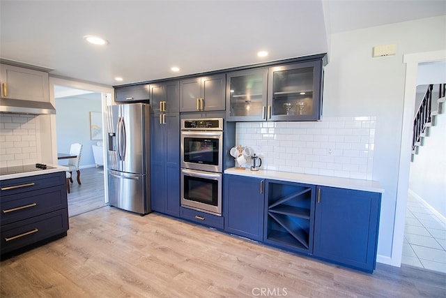 kitchen featuring blue cabinetry, range hood, backsplash, appliances with stainless steel finishes, and light wood-type flooring