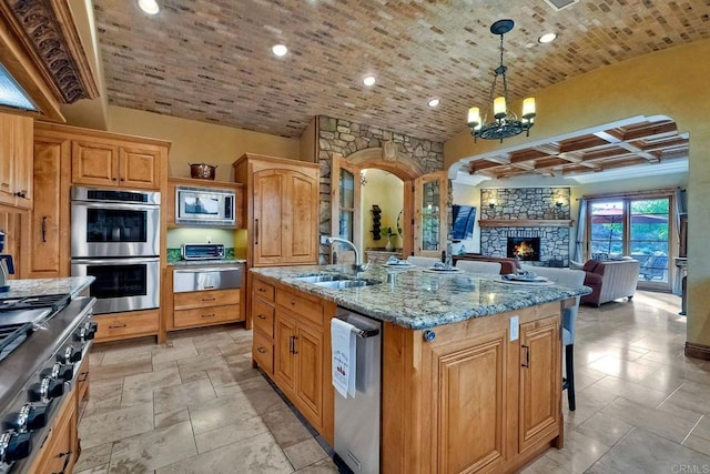 kitchen with light stone countertops, stainless steel appliances, a center island with sink, a stone fireplace, and hanging light fixtures