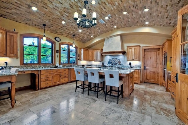 kitchen with light stone counters, premium range hood, an island with sink, vaulted ceiling, and decorative light fixtures