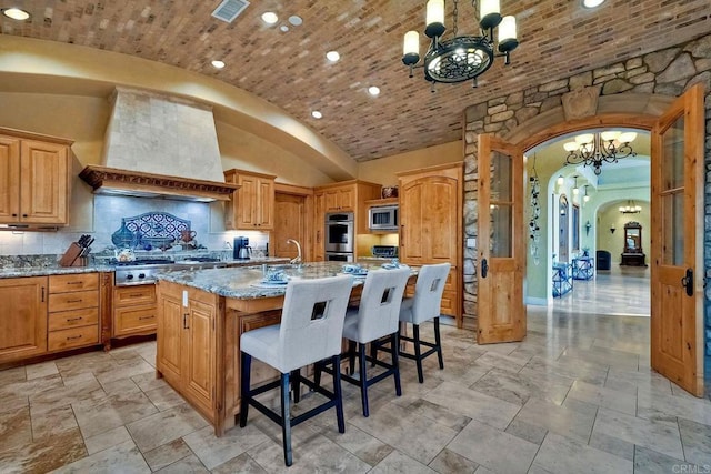 kitchen featuring light stone countertops, premium range hood, an island with sink, lofted ceiling, and appliances with stainless steel finishes