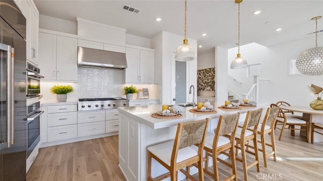 kitchen with exhaust hood, white cabinets, light wood-type flooring, an island with sink, and stainless steel appliances