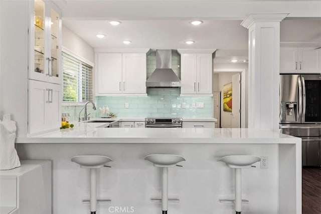 kitchen with wall chimney range hood, a breakfast bar, sink, stainless steel appliances, and white cabinets