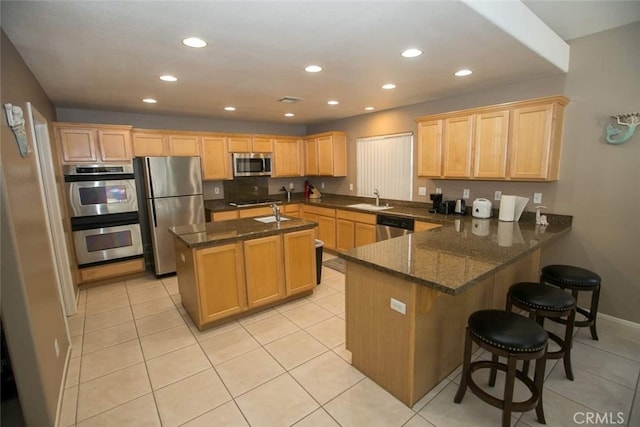 kitchen with kitchen peninsula, stainless steel appliances, a kitchen island with sink, light tile patterned floors, and dark stone countertops