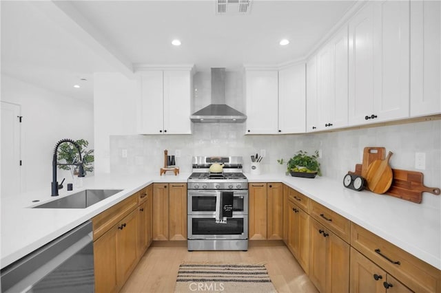 kitchen featuring dishwashing machine, wall chimney range hood, sink, white cabinets, and range with two ovens