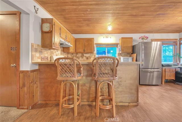 kitchen featuring white microwave, light wood-style flooring, wooden ceiling, under cabinet range hood, and freestanding refrigerator