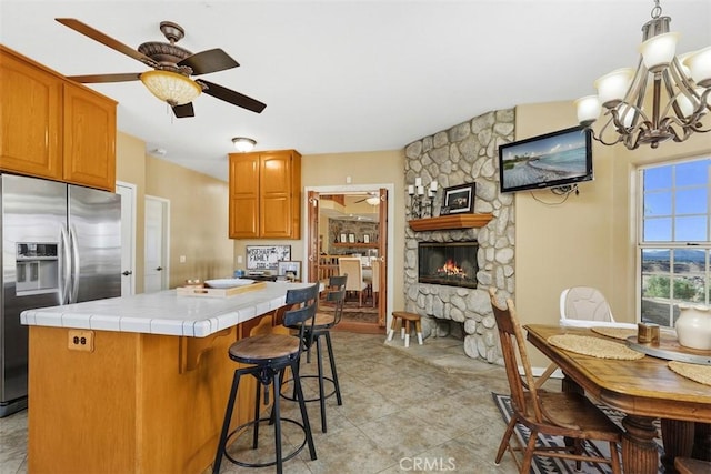 kitchen with tile counters, a fireplace, stainless steel fridge, and ceiling fan with notable chandelier